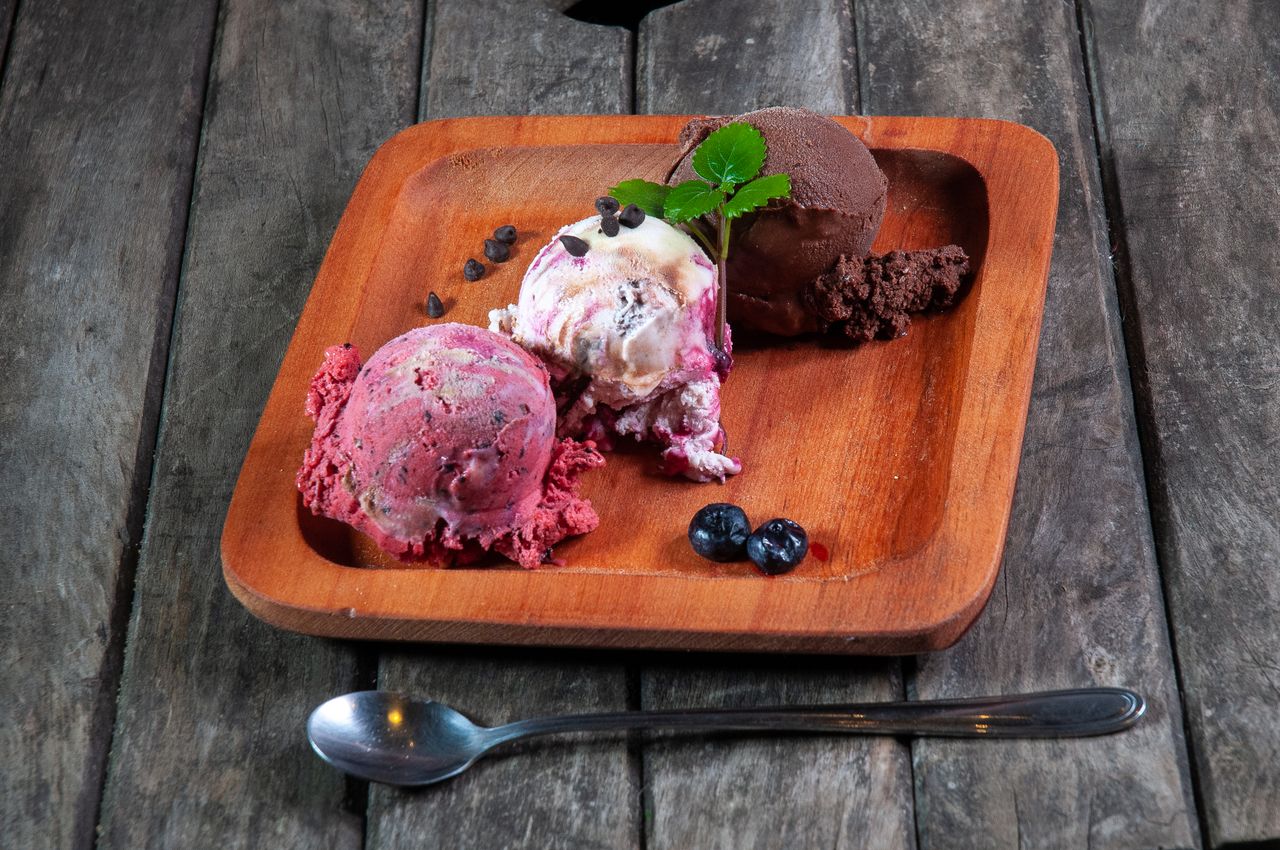 Services Three artisanal ice cream scoops on a wooden plate with chocolate chips and blueberries.