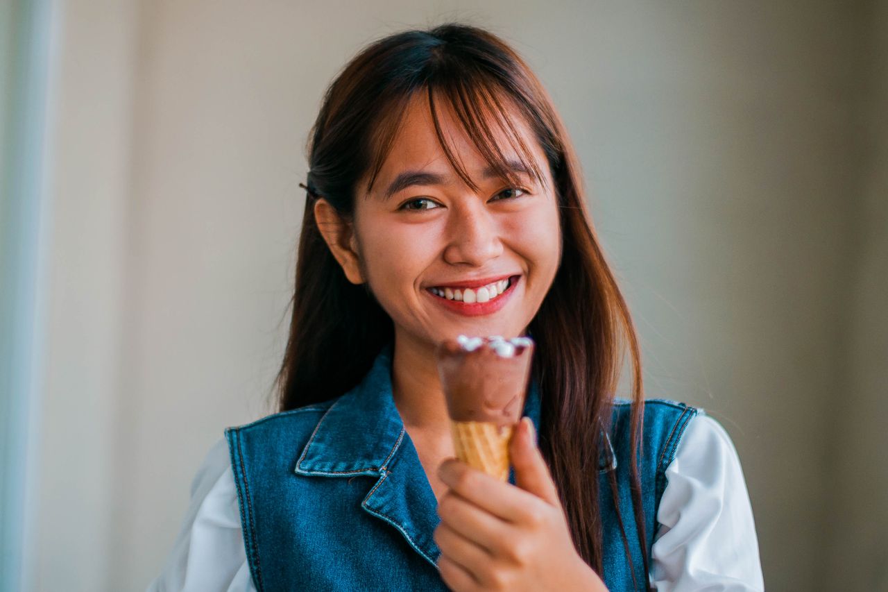 Services Positive Asian female with sweet chocolate ice cream cone in hand smiling and looking at camera while standing near wall