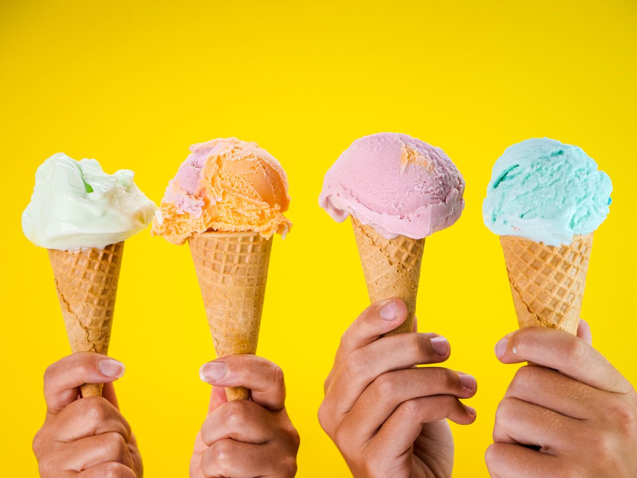 Four colorful ice cream cones held up against a bright yellow backdrop, showcasing various flavors.